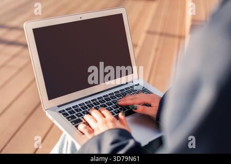 Young person using a laptop with blank screen outdoors during a sunny day on a wooden deck Stock Photo