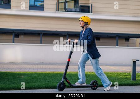 Young woman rides electric scooter on urban street wearing a yellow helmet Stock Photo