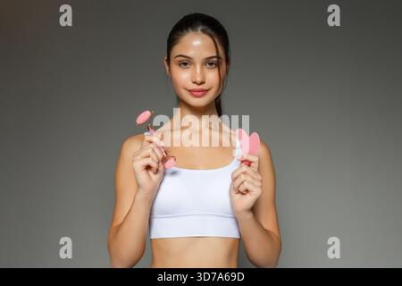 Young woman holding a pink facial roller and a gua sha stone, presenting skincare massage tools during a beauty routine on a neutral studio background Stock Photo