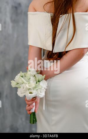 The bride in a white dress holds a wedding bouquet behind her back. Close-up of a girl in an off-the-shoulder wedding dress holding a bouquet. Stock Photo