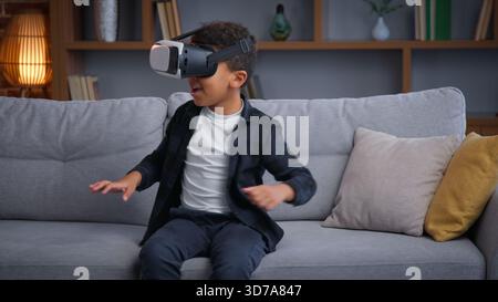 Excited African American child boy enjoying vr gaming ethnic son kid spending leisure at home with virtual reality using hobby overjoyed wearing Stock Photo