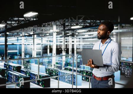 Server hub employee visualizing large language AI model using laptop, overseeing server room infrastructure from industrial platform. Data center technician reviews neural network. Stock Photo