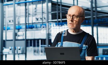 Industry 4.0 industrial plant senior engineer automating robotic machines using laptop software. Experienced technician implementing AI features in smart factory using notebook, camera A Stock Photo
