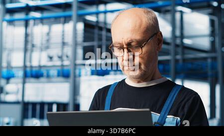 Smart factory senior engineer uses laptop to oversee operation of robotic arms and conveyor systems. Experienced industry 4.0 plant technician monitors system performance using notebook, camera A Stock Photo