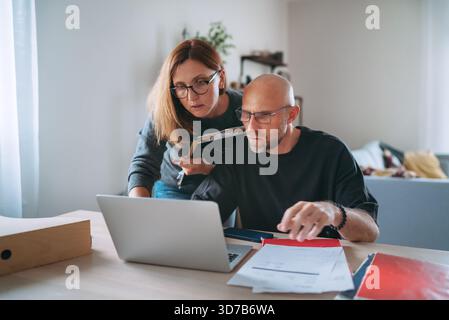Small business owners focused on financial planning at home office, reviewing paperwork and managing online tasks on laptop, showing teamwork, respons Stock Photo