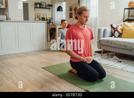 Young man kneeling on yoga mat meditating with eyes closed. Mother in background practicing breathing exercises in cozy living room for mindful relaxa Stock Photo