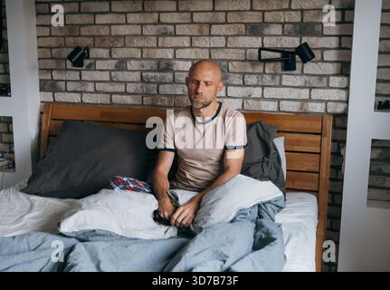 Middle-aged bald man with short beard sitting comfortably in bed during early morning wake-up moment, expressing calm mindset, stress relief, relaxati Stock Photo