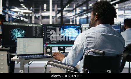 Server hub programmer with paraplegia in wheelchair uses AI machine learning node tree software. Man with disability in inclusive data center analyzes equipment issues using AI visualization, camera A Stock Photo