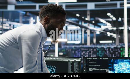 Server farm supervisor and man with disability adjust system settings based on AI analytics. Data center manager talking with technician in wheelchair overseeing automated node tree tool, camera B Stock Photo
