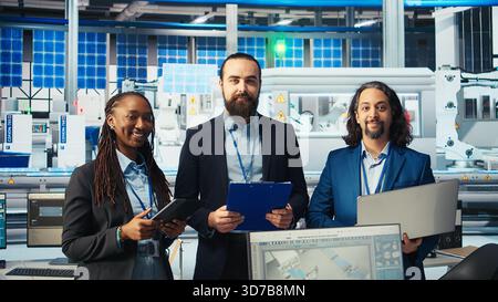 Portrait of smiling inspectors in photovoltaics factory reviewing performance of robot arms placing solar panels on assembly lines. Team of happy supervisors inspecting solar plant machinery, camera A Stock Photo