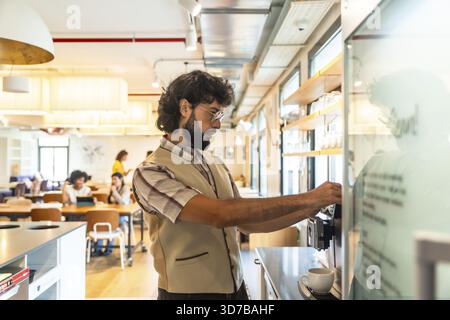 Bearded man preparing coffee at a modern coworking space, taking a relaxed break from work while colleagues chat in the bright, stylish shared office Stock Photo