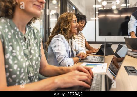 Diverse group of smiling businesswomen collaborating and engaging in productive work during a team meeting, using laptops and reviewing data in a cont Stock Photo