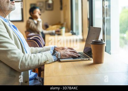 Senior professional working remotely at a busy coworking table, focused on laptop and video call with distant colleague, headset on, coffee nearby, mo Stock Photo