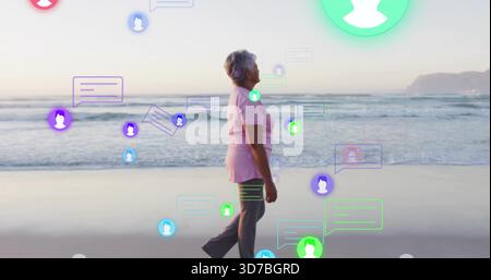 Walking senior woman in pink shirt and dark pants along sandy shoreline with colorful chat overlays Stock Photo