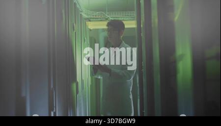 Inspecting technician in lab coat checking tablet in datacenter aisle, with server racks and LEDs Stock Photo