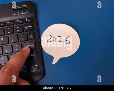 The image shows a person pressing the "Enter" key on a mini wireless Bluetooth keyboard, with a speech bubble note displaying 2026. Stock Photo
