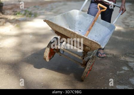 Determined worker pushing wheelbarrow with shovel at construction site. Manual labor involves moving heavy material for building, representing hard wo Stock Photo