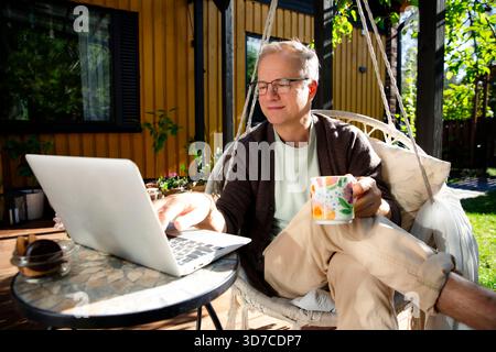 Middle-aged man working remotely from laptop and drinking coffee in hanging chair on sunny patio, relaxed lifestyle and modern home office. Stock Photo