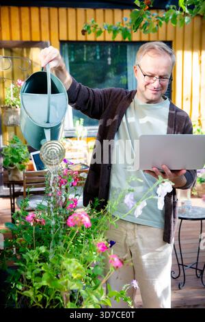 Middle-aged man watering flowers and working remotely on laptop during video call on sunny patio, modern multitasking lifestyle concept. Stock Photo