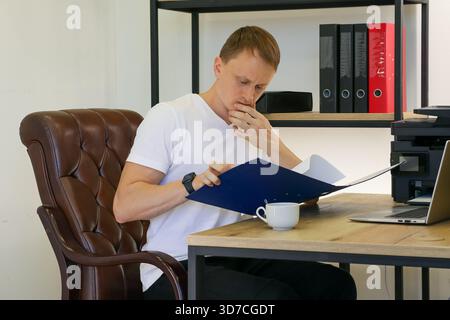 Young professional in white shirt pondering over blue folder papers in modern workspace with laptop and coffee Stock Photo
