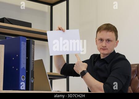 Young man in black shirt sits at desk with laptop, holds up white blank sheet in one hand while gesturing thumbs up with the other. Stock Photo
