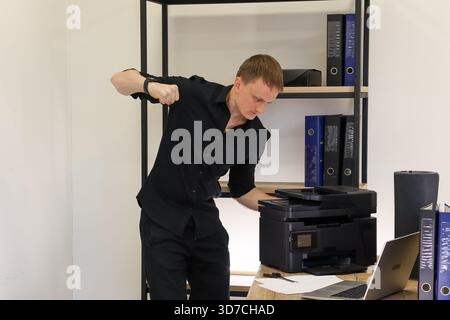 Young professional in modern office raises fist in anger at malfunctioning printer on desk with laptop and binders nearby. Stock Photo