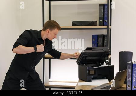 Angry man in black shirt hits jammed printer on desk with fist amid office supplies Stock Photo