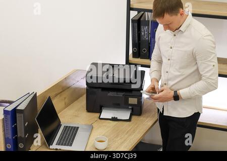 Businessman in office stands at wooden desk, holding phone near multifunction printer with laptop and files nearby. Stock Photo