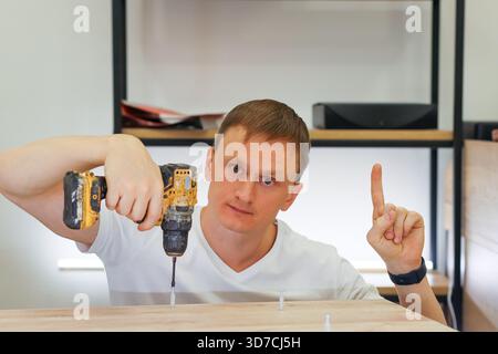 Close-up of adult male in white shirt operating yellow power drill to insert dowel into wood plank in home workshop setting. Stock Photo