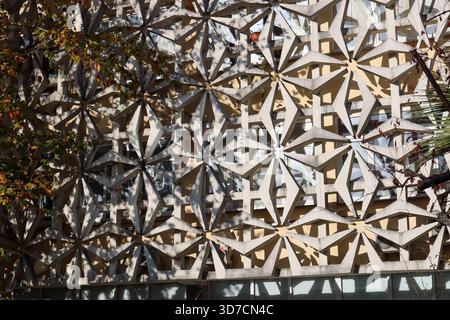 Abstract view of a modern architectural facade featuring intricate geometric concrete panels in beige tones, casting dynamic shadows amid autumn folia Stock Photo