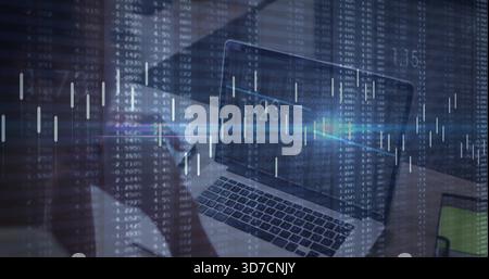 Showing laptop keys, screen at office desk, left figure wearing dark top holding phone, market data Stock Photo