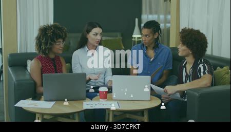 Discussing four adults blue shirt gesturing, collaborating in lounge with laptops and papers Stock Photo