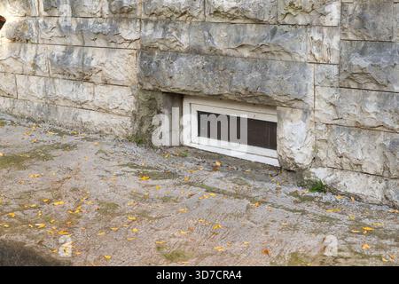 Close-up of a small basement window embedded in a rough stone wall, surrounded by scattered yellow leaves on a paved surface in autumn. Stock Photo