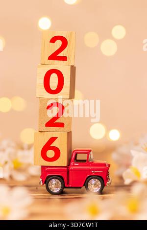 red truck toy carrying wooden cube with number 2026 in the back of the pickup for start of beginning new year 2026 with blur flower field and light Stock Photo