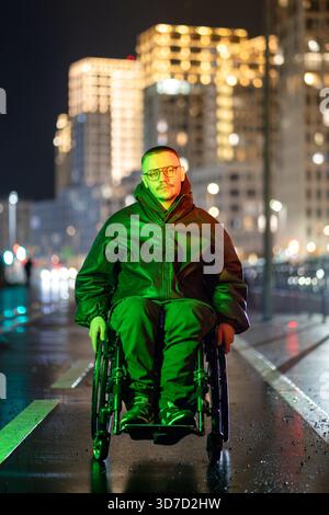 Young confident disabled man in wheelchair walking late in city, sitting in green rgb neon light. Stock Photo
