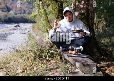 Person in white hoodie sits on log by mossy tree, raising hand in gesture while holding food container and phone beside laptop and fruits Stock Photo