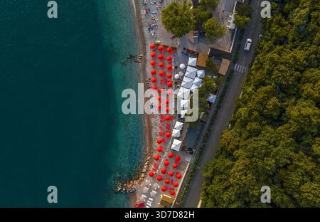 Aerial view of the Adriatic coast where striking red umbrellas line the shore against the deep blue sea, contrasting with the lush green trees, Sistia Stock Photo