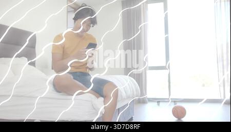 Sitting man in mustard tee, jean shorts, headset, using smartphone on bed by basketball, copy space Stock Photo