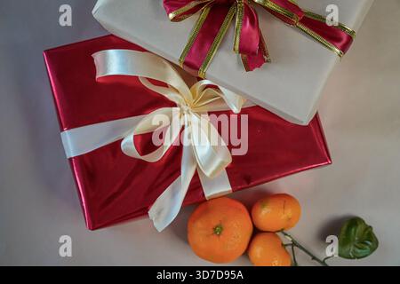 Horizontal close-up view of two stacked gift boxes, one red and one beige, decorated with ribbons. Fresh tangerines lie beside the boxes on a beige ba Stock Photo
