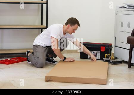 Man preparing to cut cardboard lying on floor. Adult male crouching over flat cardboard ready to use cutter knife in home workshop setting Stock Photo