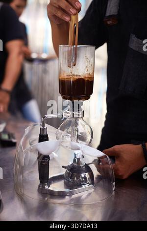 Bali, Indonesia, November 18, 2015. Close up view of man preparing coffee using siphon coffee maker, stirring grounds with wooden stick, focusing on brewing process in modern cafe Stock Photo