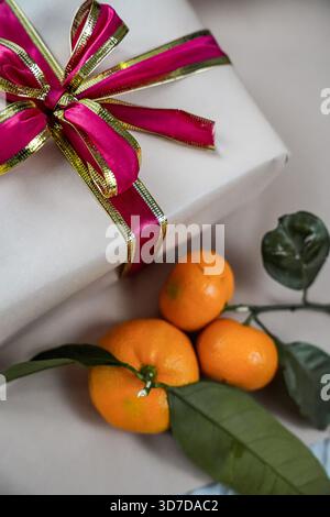Close-up of a beige gift box decorated with a red ribbon, accompanied by two fresh tangerines on a neutral background. Minimal and festive composition Stock Photo