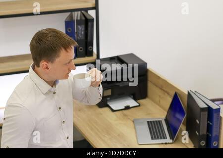 Profile of professional male consuming beverage from mug beside desk featuring laptop, printer, and binders in office interior. Stock Photo