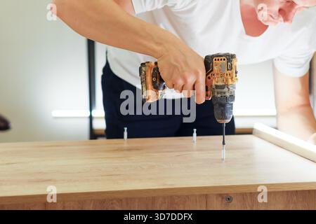 Detailed shot of person's hand holding cordless drill downward to insert fastener into light wood during furniture construction. Stock Photo