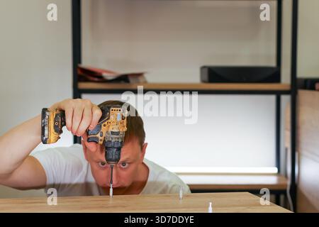 Close shot of male with wide eyes focused on inserting dowel with yellow cordless drill beneath light wood surface indoors. Stock Photo