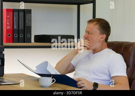 Frustrated man in casual white shirt pinches nose while examining documents in blue binder at desk with mug and computer. Stock Photo