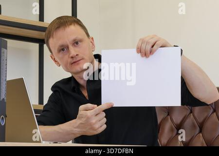 Young adult male in black shirt at desk with laptop, holding and pointing to empty white sheet for mockup purposes. Stock Photo