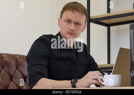 Thoughtful young businessman in formal attire holding a steaming white coffee cup at modern office desk with laptop and shelves. Stock Photo