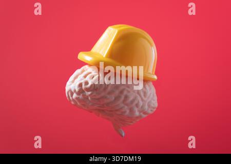 A model of a human brain wears a yellow construction hard hat against a red background. Mental protection, intellectual labor concept. Stock Photo