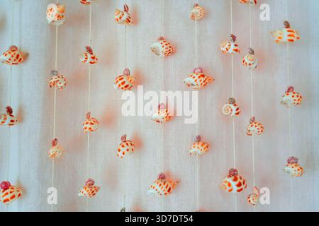 Crafted seashells with orange patterns hang elegantly from strings against a light fabric backdrop. The light illuminates the delicate decorations, cr Stock Photo